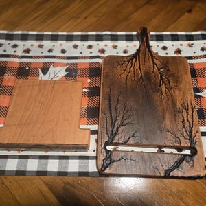 May include: Two wooden items on a patterned surface. One is a rectangular cutting board with a handle and a tree-like design. The other is a wooden phone stand. The cutting board has a slot for holding a phone or tablet.
