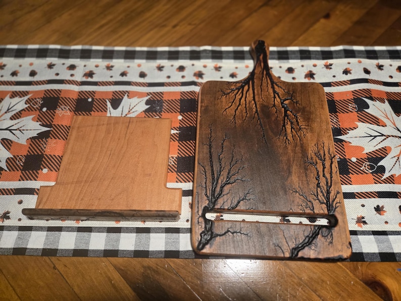 May include: Two wooden items on a patterned surface. One is a rectangular cutting board with a handle and a tree-like design. The other is a wooden phone stand. The cutting board has a slot for holding a phone or tablet.