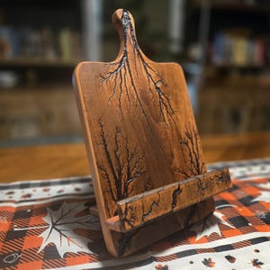 May include: Wooden cookbook stand with a handle and a built-in ledge. The stand is made of light brown wood with dark, fractal-like patterns. The stand is on a table with a fall-themed runner.
