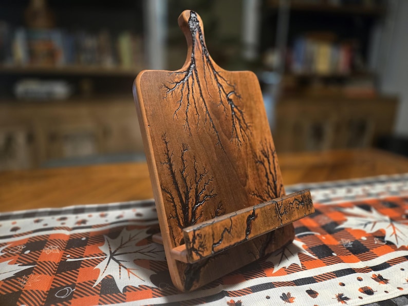 May include: Wooden cookbook stand with a handle and a built-in ledge. The stand is made of light brown wood with dark, fractal-like patterns. The stand is on a table with a fall-themed runner.
