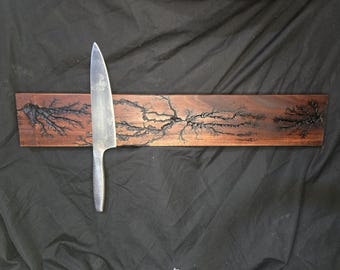 May include: A stainless steel chef's knife rests on a wooden knife holder. The holder is a rectangular piece of dark brown wood with a unique, burnt tree branch design. The knife has a textured handle and a sharp blade.