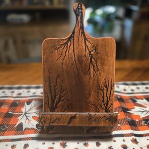 May include: A wooden cutting board with a handle and a stand. The board is a warm brown color with black fractal patterns burned into the wood. The stand is at the bottom of the board, and the handle is at the top.