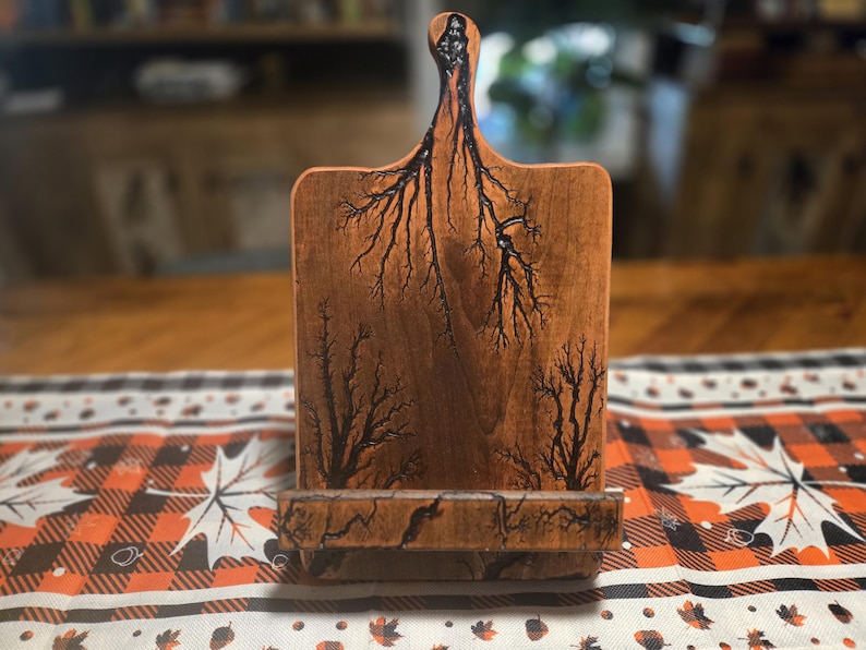 May include: A wooden cutting board with a handle and a stand. The board is a warm brown color with black fractal patterns burned into the wood. The stand is at the bottom of the board, and the handle is at the top.