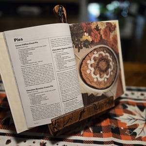 May include: A wooden cookbook stand holds open a cookbook with pie recipes. The stand is made of dark wood and has a decorative carved design. The cookbook is open to a page with a picture of a pie.
