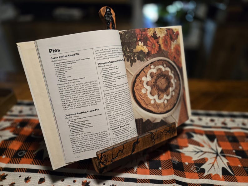 May include: A wooden cookbook stand holds open a cookbook with pie recipes. The stand is made of dark wood and has a decorative carved design. The cookbook is open to a page with a picture of a pie.