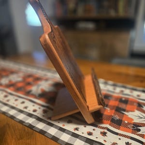 May include: A wooden cookbook stand with a natural finish. The stand is angled and features a ledge to hold a book open. The stand is on a table with a fall-themed runner.