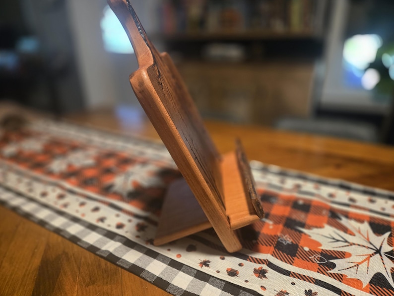 May include: A wooden cookbook stand with a natural finish. The stand is angled and features a ledge to hold a book open. The stand is on a table with a fall-themed runner.