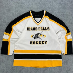 Peut inclure: Un maillot de hockey blanc, noir et jaune avec le texte IDAHO FALLS HOCKEY et un graphique d'une tête d'aigle avec des crosses de hockey croisées sur la poitrine. Le maillot à col en V a des manches noires avec des rayures jaunes et blanches.
