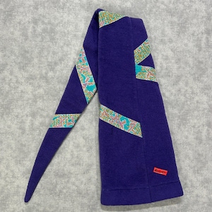 May include: A deep purple fleece scarf featuring diagonal bands of a multi-colored geometric pattern in teal, gold, and pink. A small rectangular red tag with the white text RULA'S is stitched onto the lower right corner of the fabric.