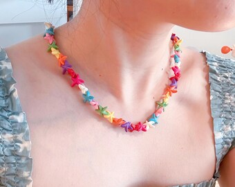 Peut inclure: Un collier coloré composé de petites perles en forme d'étoiles de différentes couleurs, notamment rose, jaune, vert et bleu. Le collier est porté autour du cou et repose sur la poitrine. La personne porte un haut à motifs bleus et blancs.
