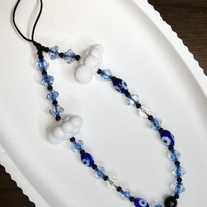 May include: A white phone charm with a black string, blue evil eye beads, and white cloud charms.