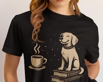 Può includere: T-shirt nero con un cane dei cartoni animati seduto su libri accanto a una tazza di caffè fumante. Il cane sorride e il disegno include piccoli puntini bianchi. La maglietta è di stile casual.