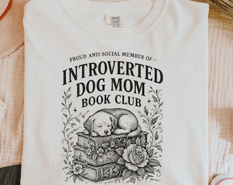 May include: Cream-colored sweatshirt with the text "PROUD ANTI-SOCIAL MEMBER OF - INTROVERTED DOG MOM BOOK CLUB." A black and white illustration of a sleeping puppy on a stack of books is below the text. The sweatshirt has a crew neck.
