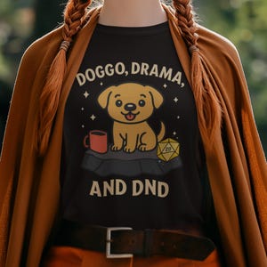 Puede incluir: Camiseta negra con el texto "Doggo, Drama, and DND". La camiseta presenta un cachorro de dibujos animados, una taza y un dado D20. El diseño es en amarillo, rojo y blanco.