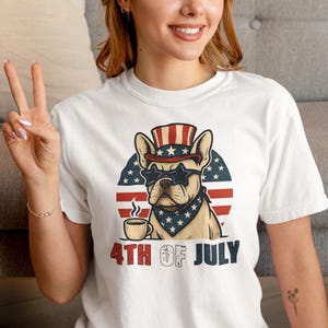 May include: White t-shirt featuring a patriotic French Bulldog wearing star-shaped sunglasses, a hat, and a bandana, with a coffee cup. The design is set against a red, white, and blue background with the text "4TH OF JULY".