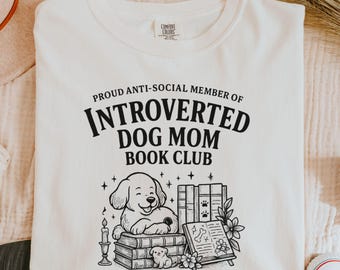 May include: Cream-colored sweatshirt with the text "PROUD ANTI-SOCIAL MEMBER OF INTROVERTED DOG MOM BOOK CLUB" in black. A graphic of a dog, books, and flowers is below the text. The sweatshirt is made of a soft material.
