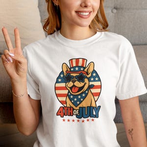 May include: White t-shirt featuring a cartoon dog wearing star-shaped sunglasses, a bandana, and a hat, all in red, white, and blue. The text "4th of July" is below the dog, with stars. The shirt is a patriotic design.
