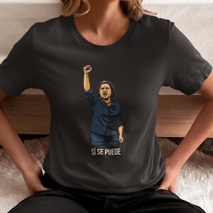 May include: Dark gray t-shirt featuring a graphic of a person with a raised fist and the words "Sí Se Puede" in white. The person in the graphic is wearing a blue shirt. The t-shirt is made of a soft material.
