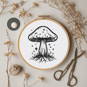 Könnte beinhalten: Ein schwarz-weißes Pilz-Kreuzstich-Design in einem hölzernen Stickrahmen. Der Pilz hat einen detaillierten Hut und Stiel mit Wurzeln und Sternen. Getrocknete Blumen und eine Vintage-Schere umgeben den Rahmen.