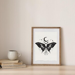 Pu&ograve; includere: Un'opera d'arte a punto croce in bianco e nero incorniciata con un disegno a farfalla con una luna crescente e stelle. L'opera d'arte &egrave; in una cornice di legno e poggia su una superficie di legno. Sono visibili anche una pila di libri e una tazza.