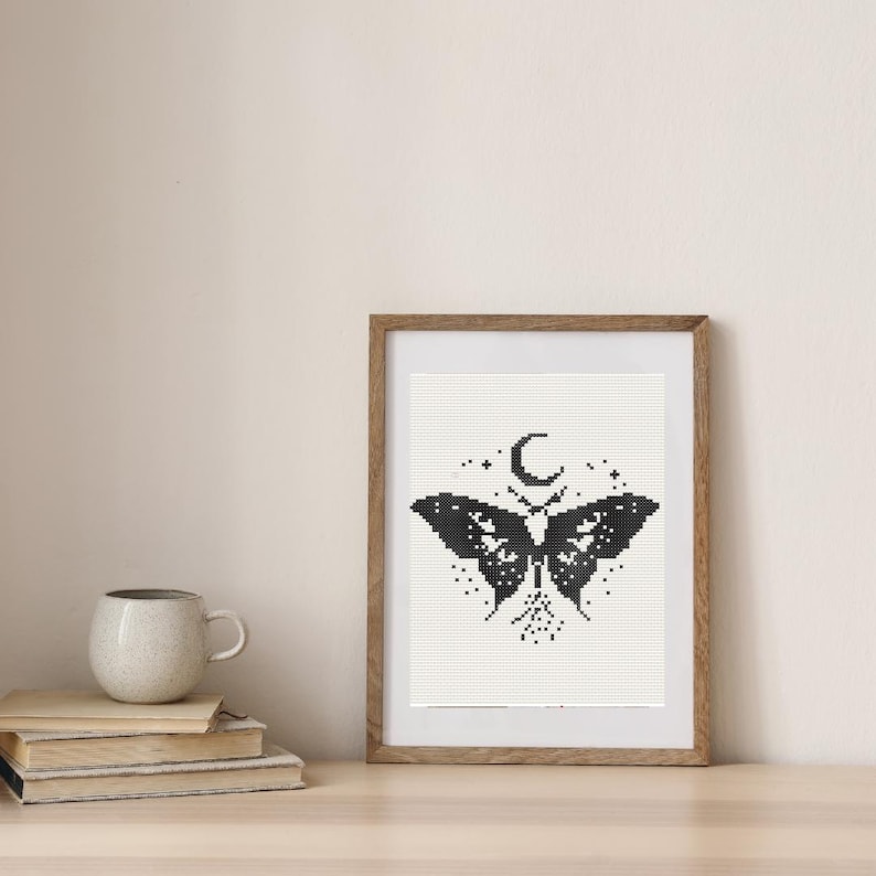 Pu&ograve; includere: Un'opera d'arte a punto croce in bianco e nero incorniciata con un disegno a farfalla con una luna crescente e stelle. L'opera d'arte &egrave; in una cornice di legno e poggia su una superficie di legno. Sono visibili anche una pila di libri e una tazza.