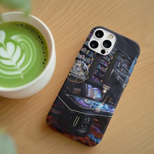 Op de afbeelding: Zwarte smartphonehoes met een gedetailleerde illustratie van een bibliotheekinterieur. Een kopje groene matcha latte met latte art staat naast de telefoon op een houten oppervlak.