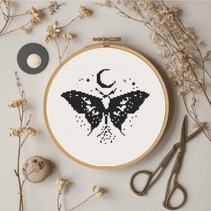 May include: A cross-stitch embroidery in a wooden hoop features a black moth design with a crescent moon and stars. The artwork is surrounded by dried flowers, a gray circle, and vintage scissors, creating a rustic, handcrafted aesthetic.