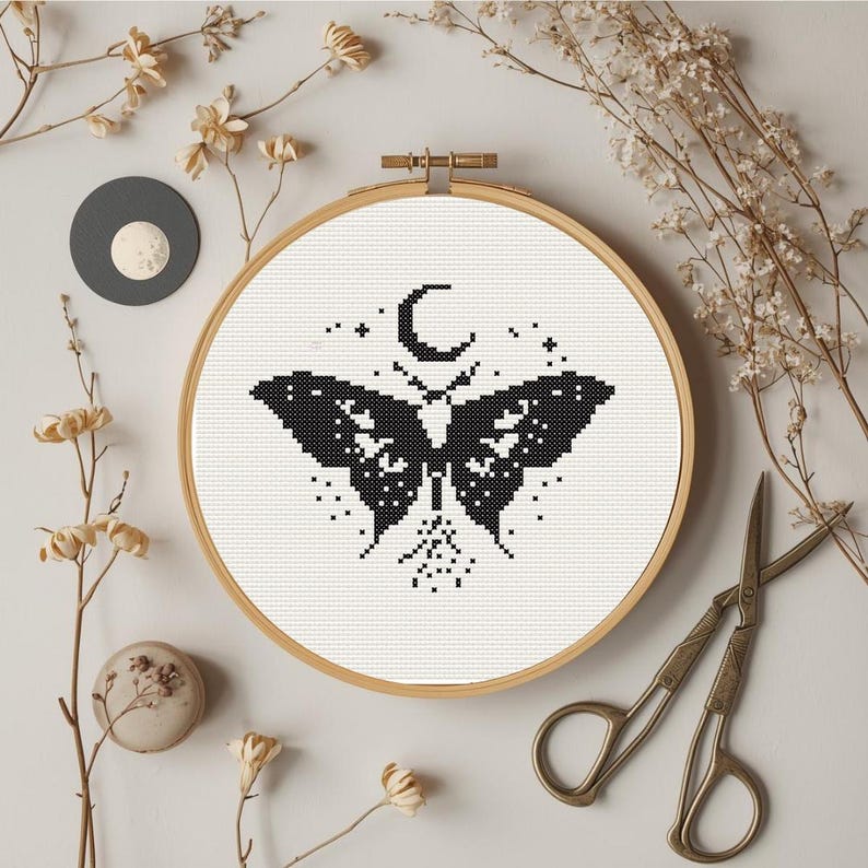 Pu&ograve; includere: Ricamo a punto croce in un telaio di legno con un disegno di falena nera con luna crescente e stelle. L'opera &egrave; circondata da fiori secchi, un cerchio grigio e forbici vintage, creando un'estetica rustica e artigianale.