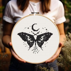 Pu&ograve; includere: Un disegno a punto croce in bianco e nero di una falena con una luna crescente e stelle, esposto in un telaio da ricamo in legno. L'opera &egrave; tenuta da una persona.
