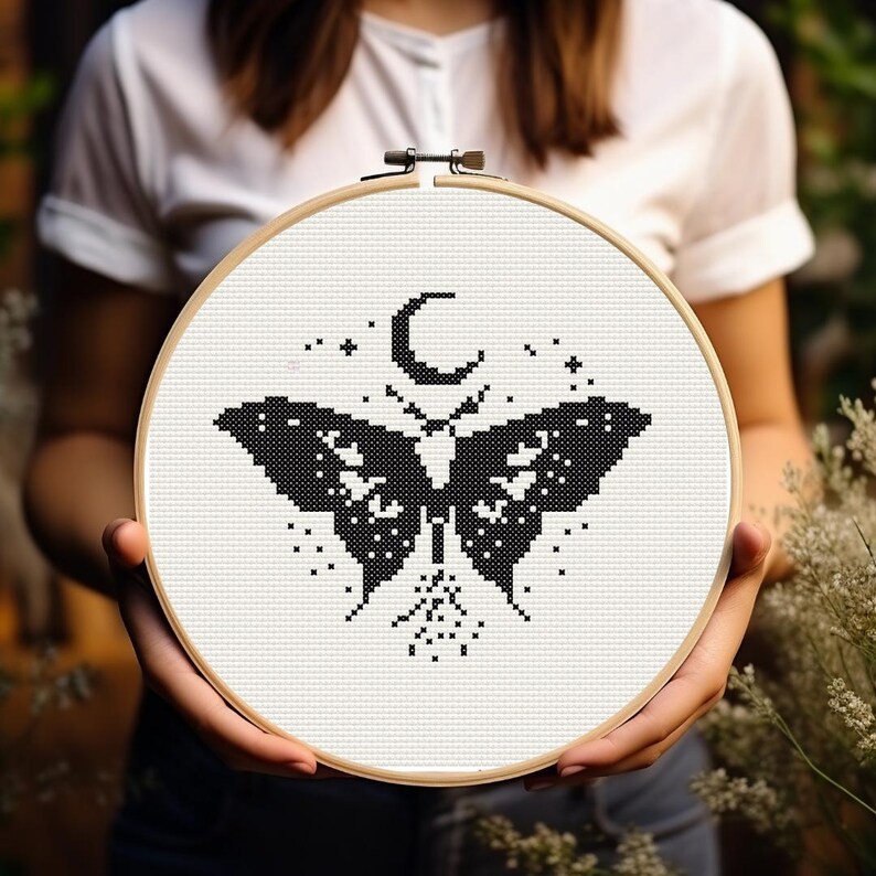 Pu&ograve; includere: Un disegno a punto croce in bianco e nero di una falena con una luna crescente e stelle, esposto in un telaio da ricamo in legno. L'opera &egrave; tenuta da una persona.