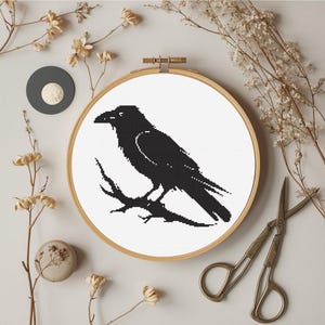 May include: A black crow cross-stitch design in a wooden embroidery hoop. The crow is perched on a branch against a white background. Dried flowers and vintage scissors surround the hoop, creating a rustic, handcrafted aesthetic.
