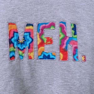 May include: A gray sweatshirt with the word "MEH" embroidered in colorful tie-dye letters. The letters are outlined with a contrasting thread, and the overall design has a casual, artistic feel.