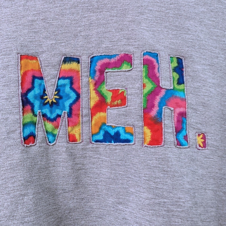 May include: A gray sweatshirt with the word "MEH" embroidered in colorful tie-dye letters. The letters are outlined with a contrasting thread, and the overall design has a casual, artistic feel.