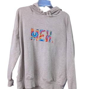 May include: A light gray hoodie with a drawstring hood and long sleeves. The word "MEH" is embroidered across the chest in a colorful, tie-dye style font. The hoodie is hanging on a blue hanger.