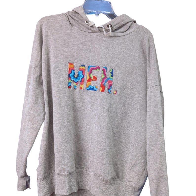 May include: A light gray hoodie with a drawstring hood and long sleeves. The word "MEH" is embroidered across the chest in a colorful, tie-dye style font. The hoodie is hanging on a blue hanger.