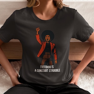 Puede incluir: Camiseta gris oscuro con un gráfico de una mujer con el puño en alto, con una chaqueta roja. El texto "FREEDOM IS A CONSTANT STRUGGLE" está impreso en blanco. La camiseta es de cuello redondo.