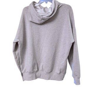 May include: A light gray hoodie with a drawstring hood and long sleeves. The hoodie is hanging on a clear plastic hanger. The interior of the hood is white, and there is a light blue accent on the sleeve.