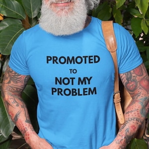 Pode incluir: Uma camiseta azul clara com o texto "PROMOTED TO NOT MY PROBLEM" em preto. A camiseta é usada por uma pessoa com barba branca e tatuagens nos braços.