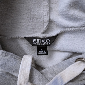 May include: Close-up of a gray hoodie with a drawstring. The black label reads "BUFFALO DAVID BITTON" and "L MADE IN CAMBODIA". The hoodie has white drawstrings and metal eyelets.