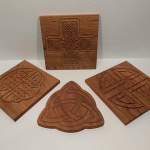 May include: A selection of wooden carvings with Celtic knot patterns. The carvings are in different shapes, including squares and a triangle, and are made from a warm, reddish-brown wood. The designs are intricately carved into the wood.