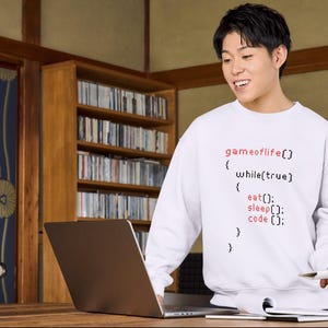 Peut inclure: Sweat-shirt blanc avec le texte "gameoflife()" et des extraits de code en rouge et noir. La personne tient un stylo et un ordinateur portable est sur le bureau. L'arrière-plan comprend une bibliothèque en bois et une pièce de style japonais traditionnel.