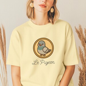 May include: A light yellow t-shirt featuring a cartoon pigeon inside a gold-colored circle. The pigeon is gray, teal, and pink. The text "Le Pigeon" is written below the circle in a cursive font.