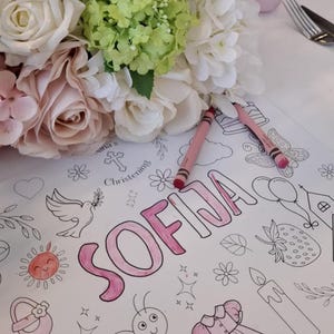 May include: A coloring page with the name "SOFIA" in pink, surrounded by line drawings of a dove, sun, flowers, and a dress. Two pink crayons are on the page. A bouquet of white, pink, and green flowers is in the background.