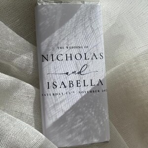 May include: A white rectangular wedding favor with the text "THE WEDDING OF NICHOLAS and ISABELLA SATURDAY 15TH NOVEMBER 202" in black. The favor is wrapped in clear cellophane, with shadows cast across the surface.