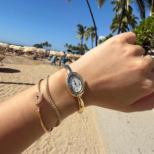 May include: A gold-toned watch with a white face and Roman numerals, paired with two gold bracelets. One bracelet features a heart-shaped design with small stones. The other is a delicate chain. The watch and bracelets are on a wrist.