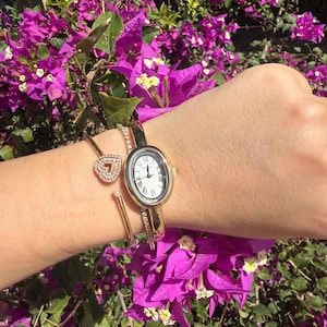 May include: A gold-toned watch with a white face and Roman numerals, paired with a gold heart-shaped bracelet. The watch has a slender band adorned with small crystals. The jewelry is displayed against a backdrop of vibrant purple flowers.