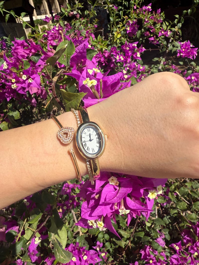 May include: A gold-toned watch with a white face and Roman numerals, paired with a gold heart-shaped bracelet. The watch has a slender band adorned with small crystals. The jewelry is displayed against a backdrop of vibrant purple flowers.