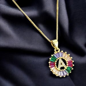 May include: A gold chain necklace with a pendant featuring the letter "A" surrounded by colorful gemstones. The pendant is round and has a polished finish.