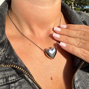 May include: A silver heart-shaped pendant necklace on a silver chain. The necklace is worn by a person with a light complexion and is shown against a dark denim background.