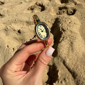 May include: Gold-toned oval wristwatch with a cream-colored face and Roman numeral hour markers. The watch has a gold band and is held against a sandy background. The watch face displays the time.